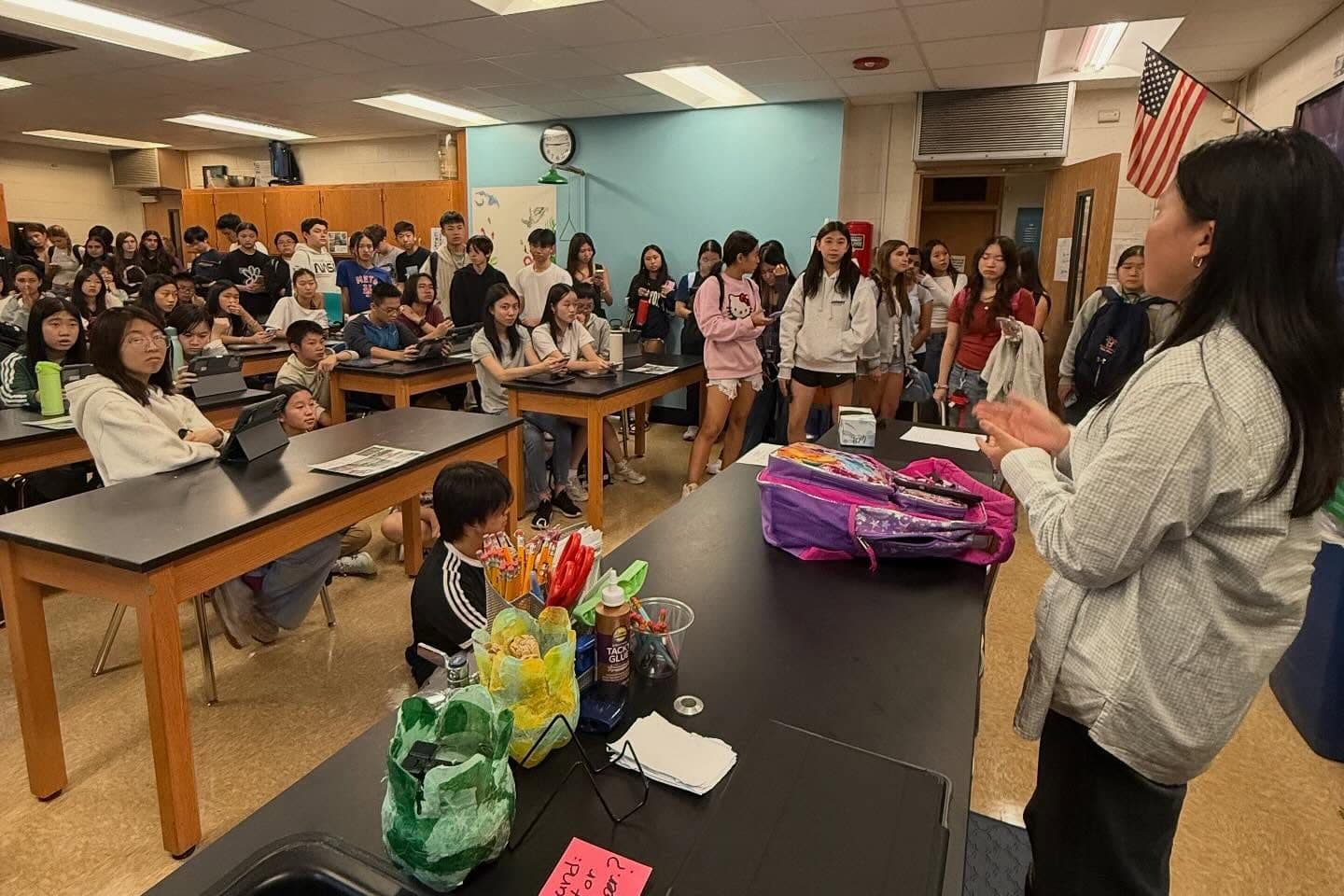 A science classroom packed with students at a club meeting. Around forty sit at black-topped lab benches or stand along the back wall, most facing right towards an adult in a pale checked shirt who is speaking with her hands raised. A small flag and a wall clock hang above.