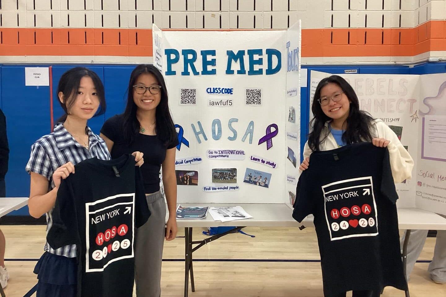 Three students stand behind a table in a school gymnasium, two of them holding up black T-shirts lettered NEW YORK HOSA. Behind them a hand-lettered tri-fold display board reads PRE MED and, below that, HOSA, with a blue and a purple awareness ribbon and small photographs pinned around it. Other clubs' tables are set up on either side.