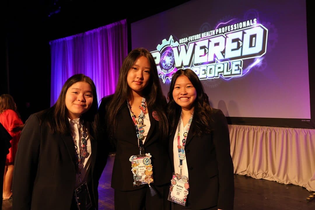 Three students in dark business suits stand together on a stage, each wearing a conference lanyard hung with enamel pins and badges. Behind them a large screen reads HOSA - Future Health Professionals, Powered by People, against a purple-lit curtain.