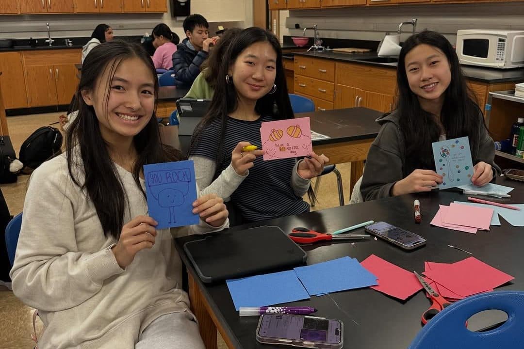 Three students sit along a black lab bench, smiling and holding up hand-drawn cards - one blue, one pink, one pale blue. Sheets of coloured paper, markers and scissors are spread across the bench in front of them, and more students work at benches behind.