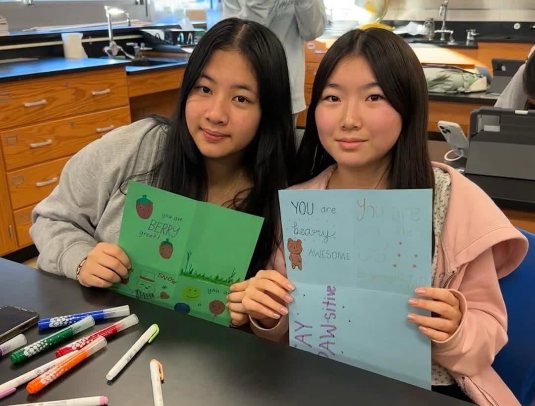 Two students sit at a black lab bench holding open hand-made cards towards the camera. One card is green with drawn strawberries and the words you are BERRY great; the other is pale blue with a drawn bear and the words you are beary awesome. Felt-tip pens are scattered on the bench in front of them.
