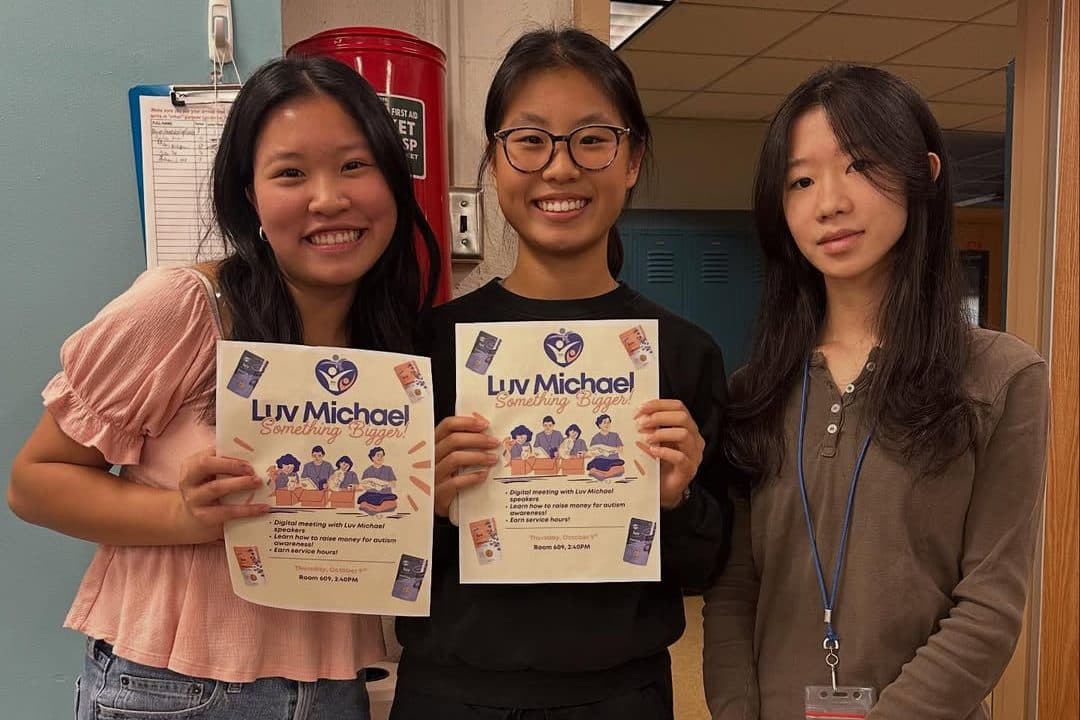 Three students stand side by side in a school corridor in front of a red first-aid cabinet and blue lockers. Two of them hold up printed flyers announcing a club session run with a volunteering organisation; the third stands with her hands clasped, wearing a lanyard.